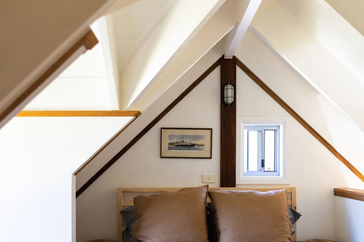 Un dormitorio con techo inclinado, una cama con almohadas, una mesita y un ventanal.