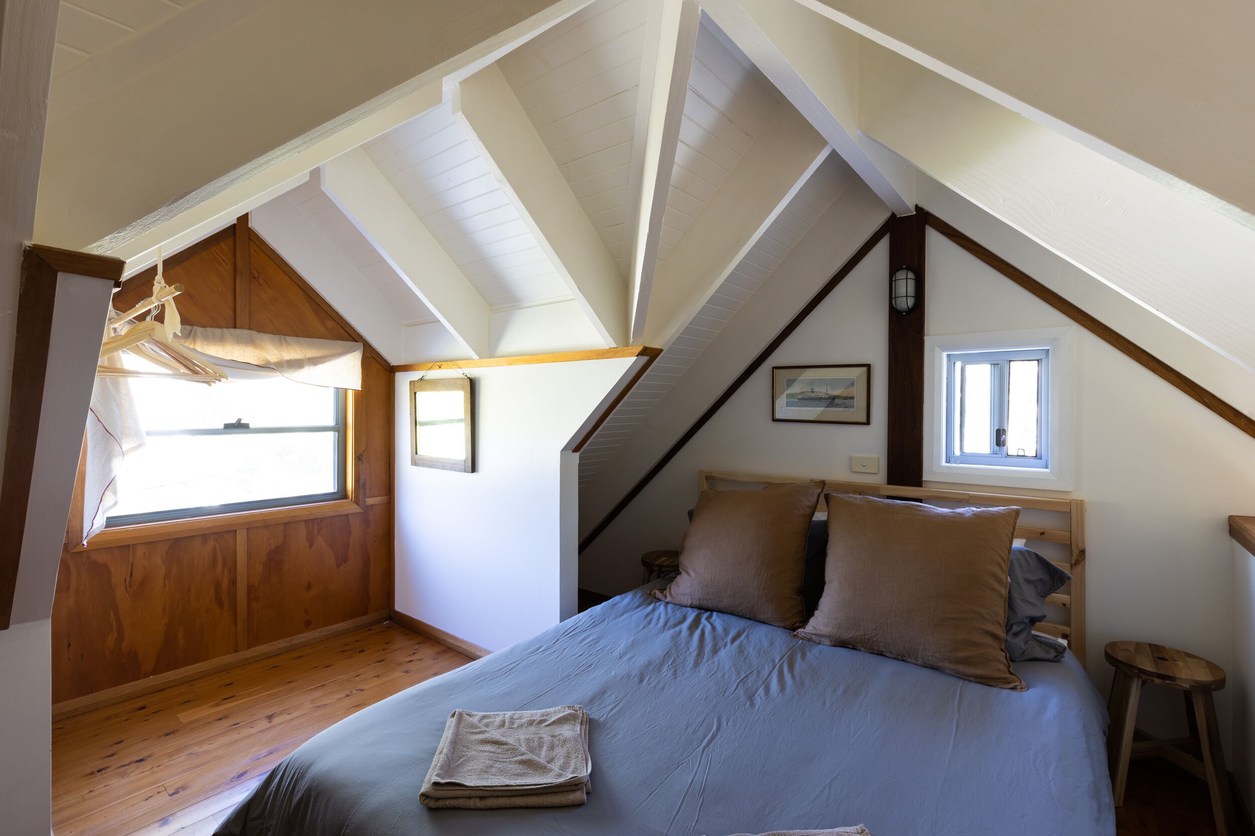 A bedroom with a sloped ceiling, wooden beams, a bed with blue bedding, and a window providing natural light.