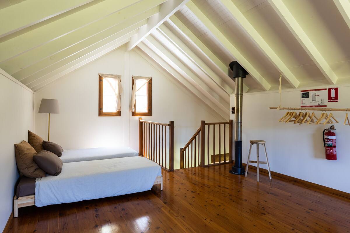 Un dormitorio con cama, suelo de madera, una escalera, un extintor y una ventana.