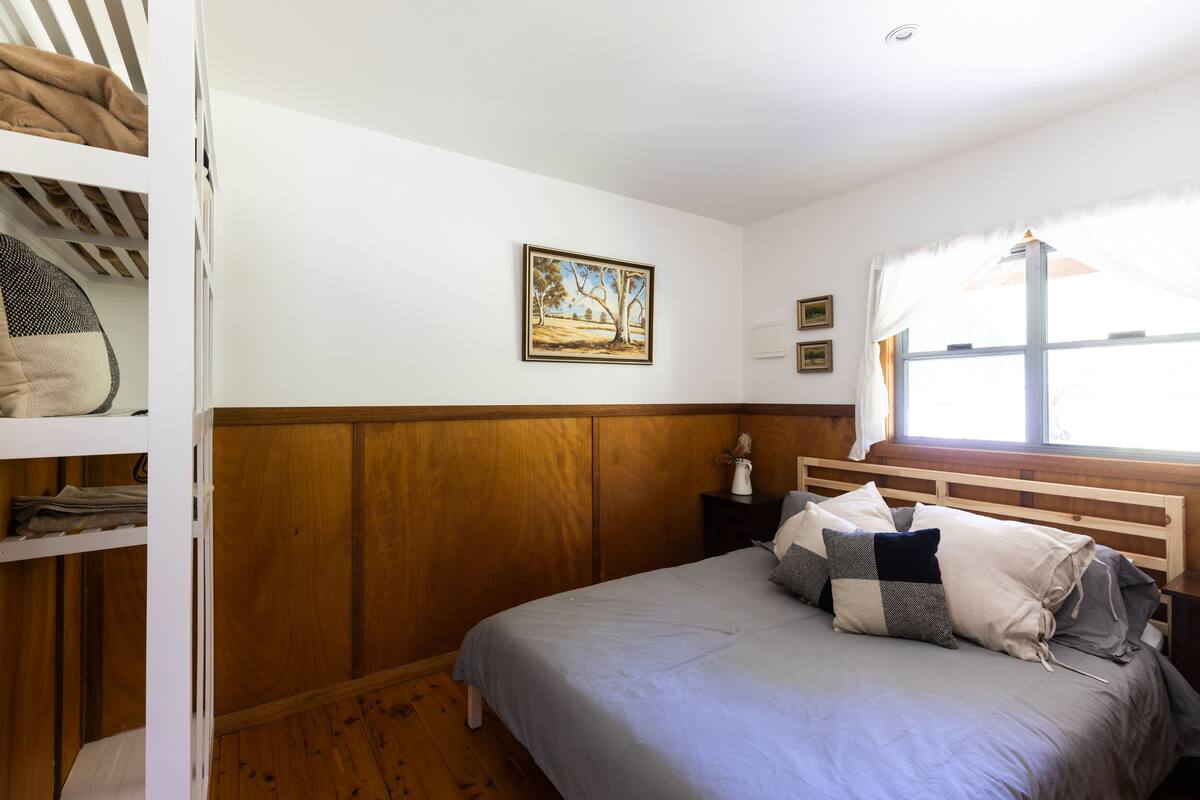 Un dormitorio con cabecera de madera, cama con ropa de cama gris, estante blanco con ropa doblada, un cuadro enmarcado de paisaje y una ventana con cortinas translúcidas.