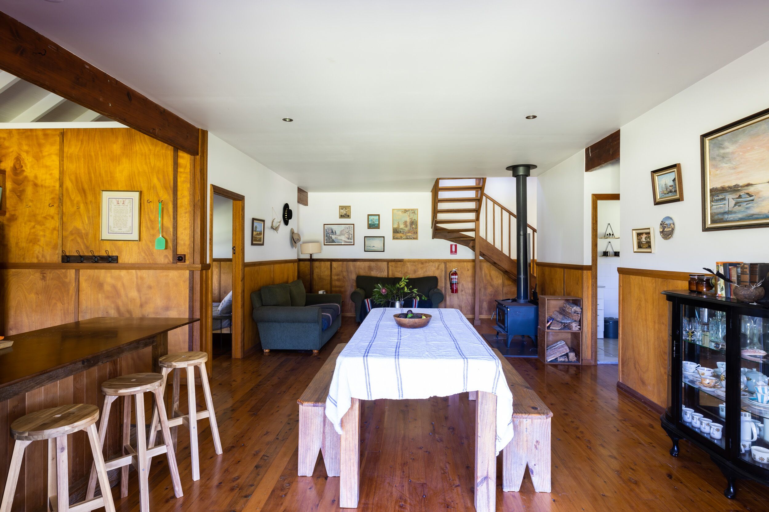 A wooden-floored room with a dining table, bar stools, a staircase, and a cabinet with glassware.