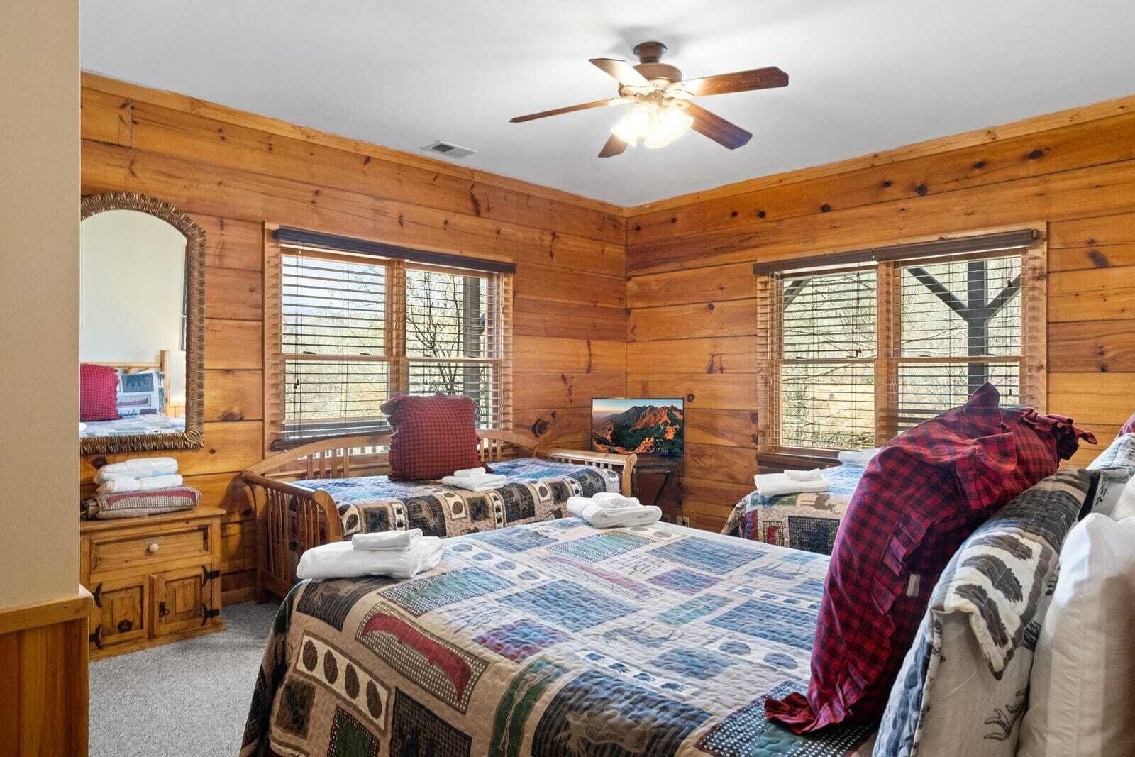 Un dormitorio estilo cabaña con dos camas, un ventilador de techo de madera y ventanas amplias.