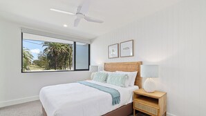 Un dormitorio con una cama grande, una mesita de noche con lámpara, un ventilador de techo y una ventana con vistas a palmeras.