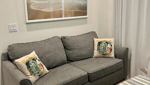 Un divano grigio con due cuscini decorativi, un quadro raffigurante una spiaggia e un poggiapiedi a righe.