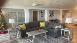 Een woonkamer met een grijze modulaire bank, twee bijzettafels met lampen, een salontafel, een kerstboom en een keuken op de achtergrond.