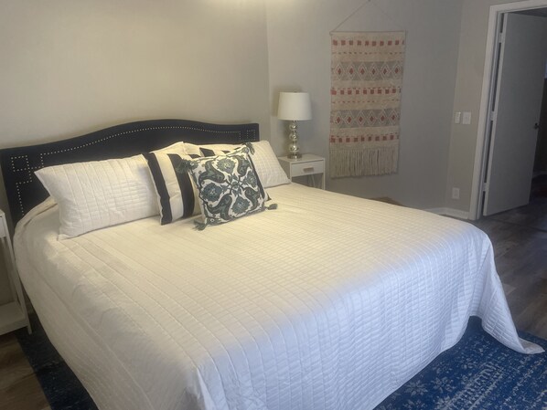 Un lit bien fait, avec une literie blanche et des coussins décoratifs, une table de chevet avec une lampe, et une tapisserie à motifs accrochée au mur.