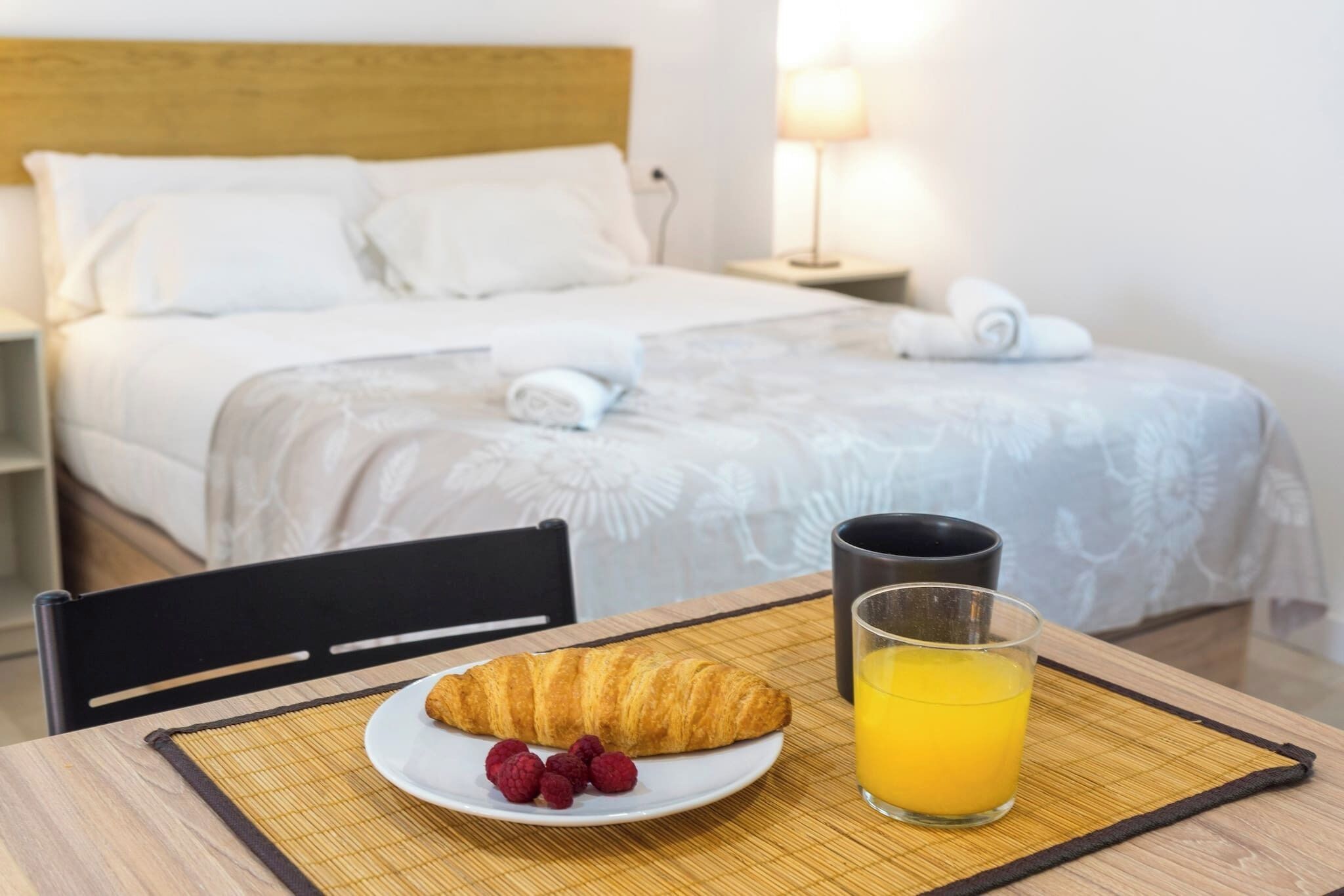 A hotel room with a neatly made bed, a breakfast table with croissant, raspberries, and orange juice, and a bedside lamp.