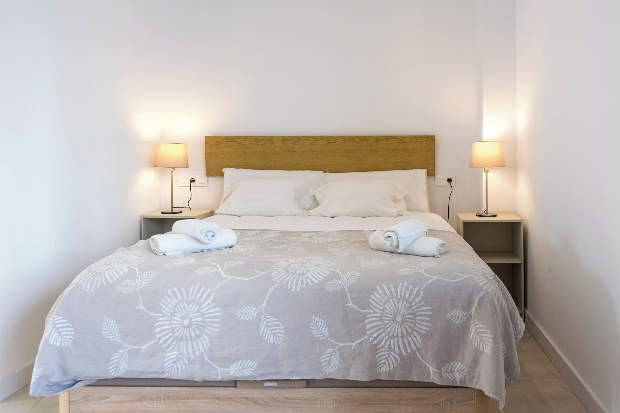 Una cama bien hecha con una colcha estampada de flores, dos lámparas de noche y un cabecero de madera.