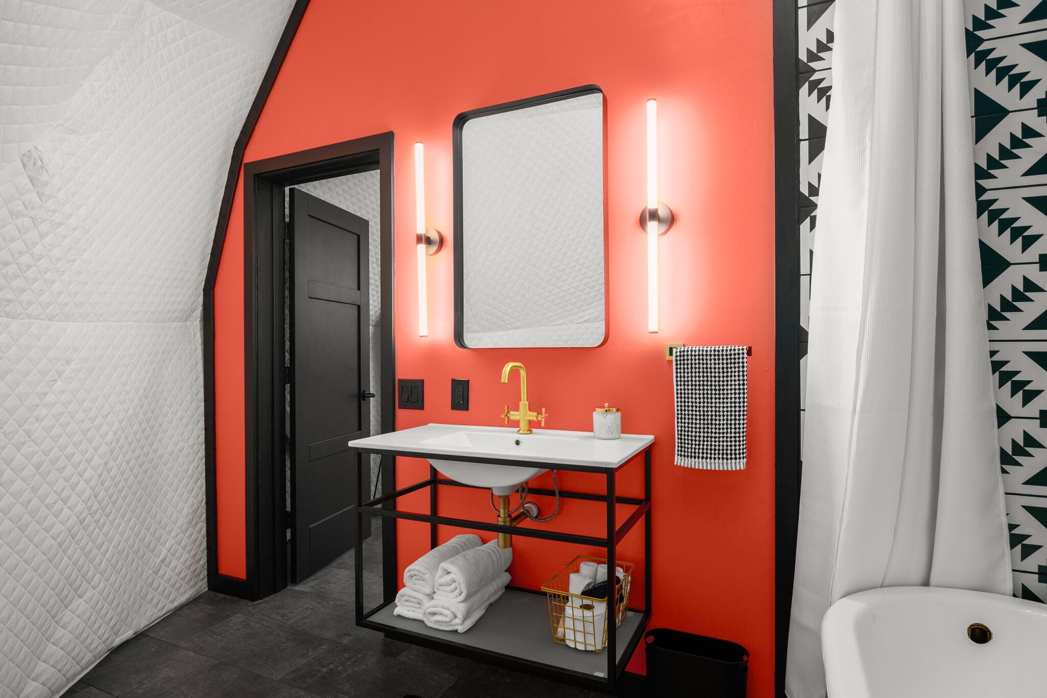 Ein Badezimmer mit einem weißen Waschbecken, einem Spiegel und einer roten Wand. Handtuchhalter mit gefalteten Handtüchern und eine Mülleimfach sind ebenfalls vorhanden.