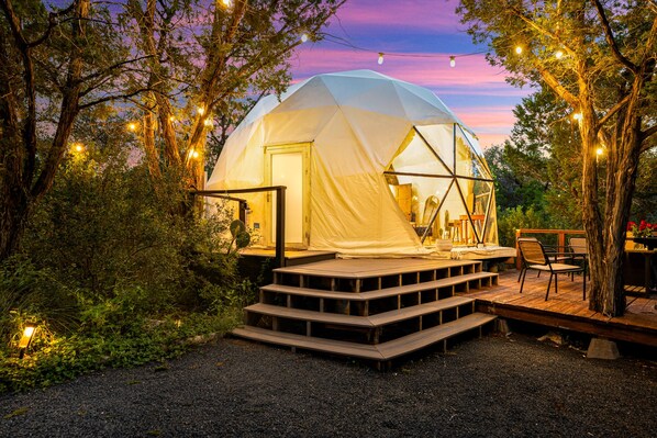 A geodesic dome tent with a wooden deck and outdoor seating area, surrounded by trees and string lights. - Hill Country Glamping Dome w/Hot Tub, Gym, Sauna & Cold Plunge (Spicewood)