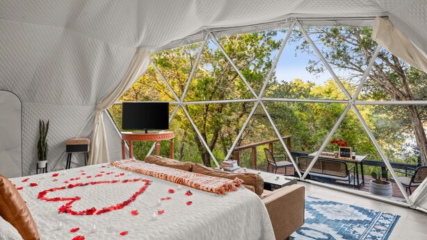 Uma tenda espaçosa e bem iluminada, com uma cama decorada com pétalas de rosas vermelhas, televisão e área de refeições com vista para as árvores.