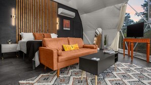 Interior moderno com cama, sofá e área de televisão, apresentando um teto com design diferenciado em forma de tenda e vista para árvores.