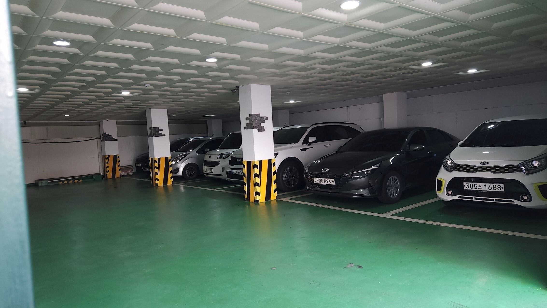 Un parking souterrain où plusieurs voitures sont garées dans des emplacements prévus à cet effet, avec des piliers blancs rayés de jaune et un sol vert.