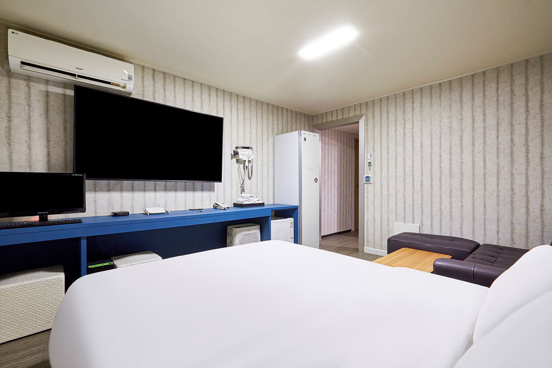 Una camera d'albergo con un letto, una scrivania con un computer, una televisione, un frigorifero e un piccolo angolo salotto.