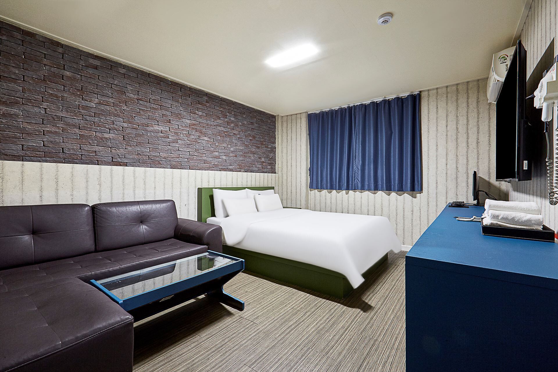 A hotel room with a brick accent wall, a bed with white linens, a dark leather sofa, a desk with a telephone, and a window with blue curtains.