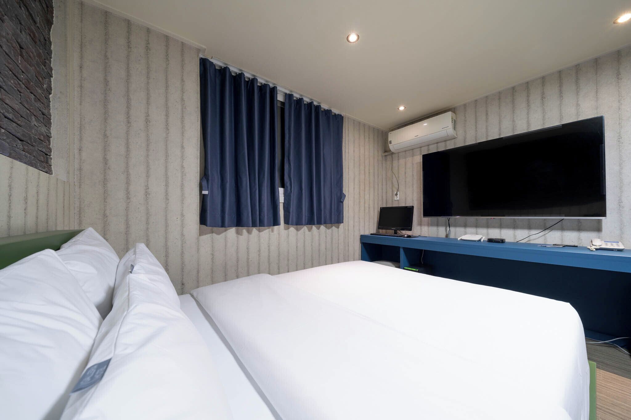 A hotel room with a bed, a desk with a computer, a flat-screen TV, and an air conditioning unit.
