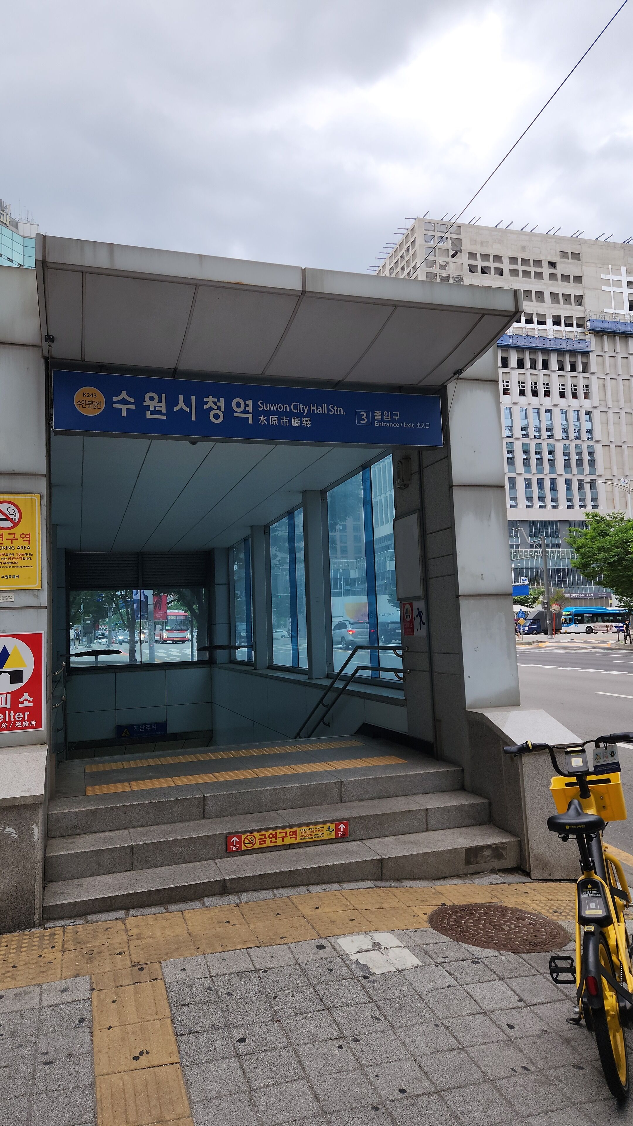 Une entrée d’immeuble avec une enseigne bleue indiquant la station de métro « Suwon City Hall » et une rampe d’accès pour les personnes à mobilité réduite.