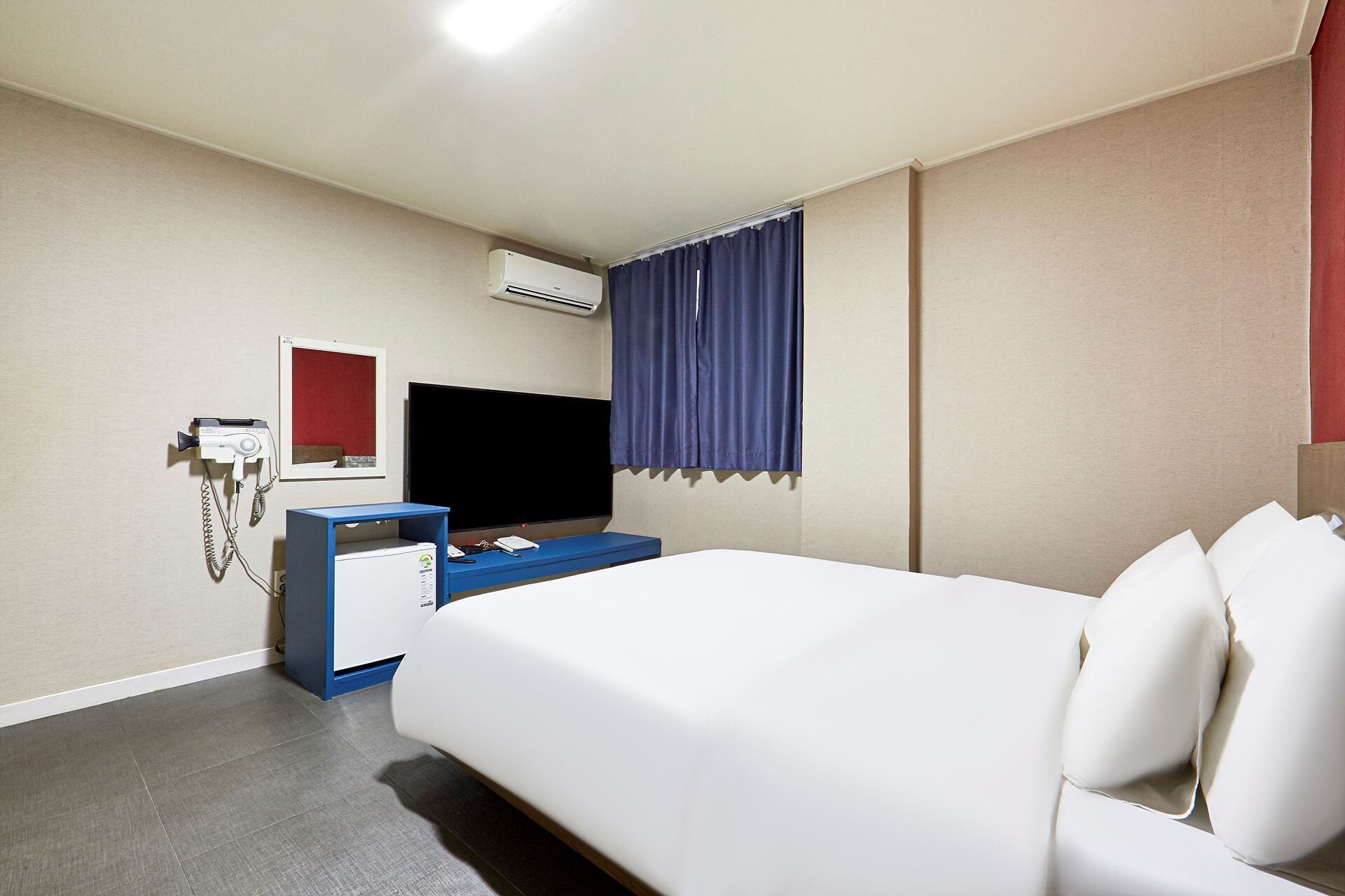 A hotel room with a bed, a desk with a flat-screen TV, an air conditioning unit, and a telephone.