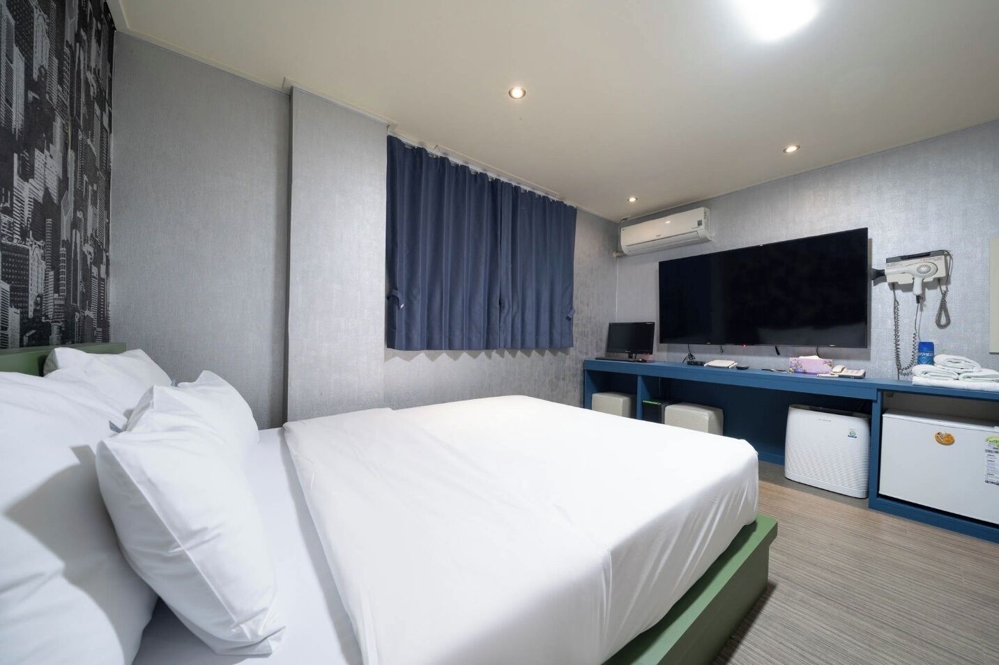 A hotel room with a large bed, a desk with a computer, a flat-screen TV, an air conditioning unit, and a telephone.