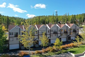 A row of two-story townhouses with a garage on the ground floor, surrounded by trees and a forested hillside. - The Powderhound Condo (Whitefish)
