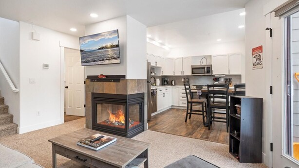 A modern living room with a fireplace, a dining area, and a kitchen with white cabinets and stainless steel appliances. - The Powderhound Condo (Whitefish)
