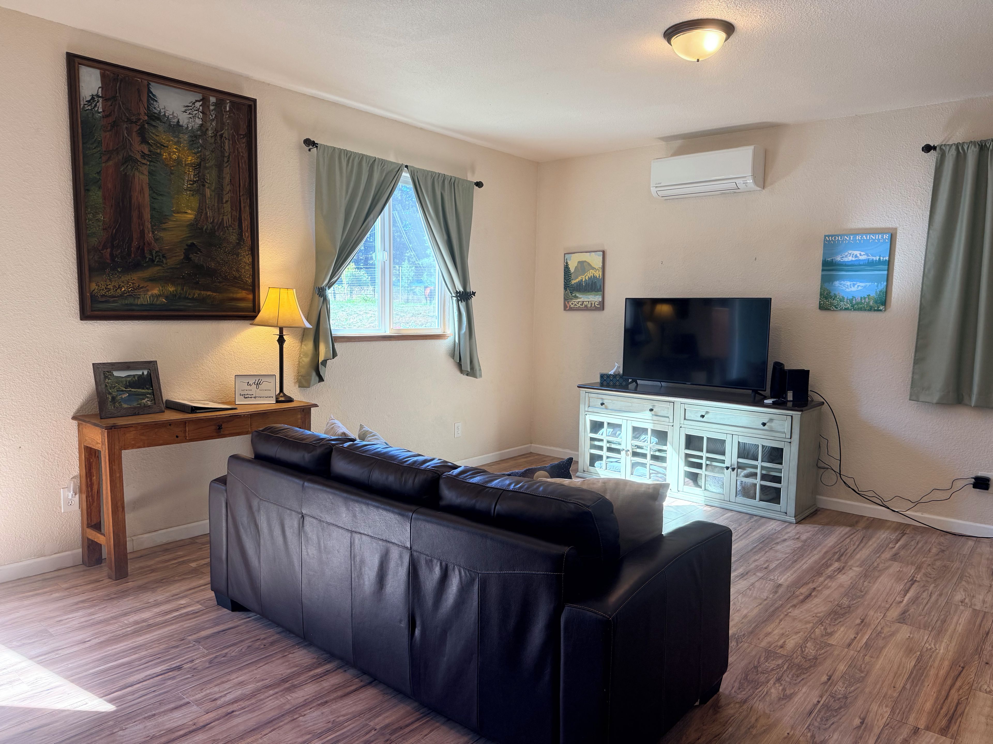 Ein Wohnzimmer mit einem schwarzen Ledersofa, einem Holztisch mit Lampe, einem Fernseher auf einer weißen Kommode und einem Bild an der Wand.