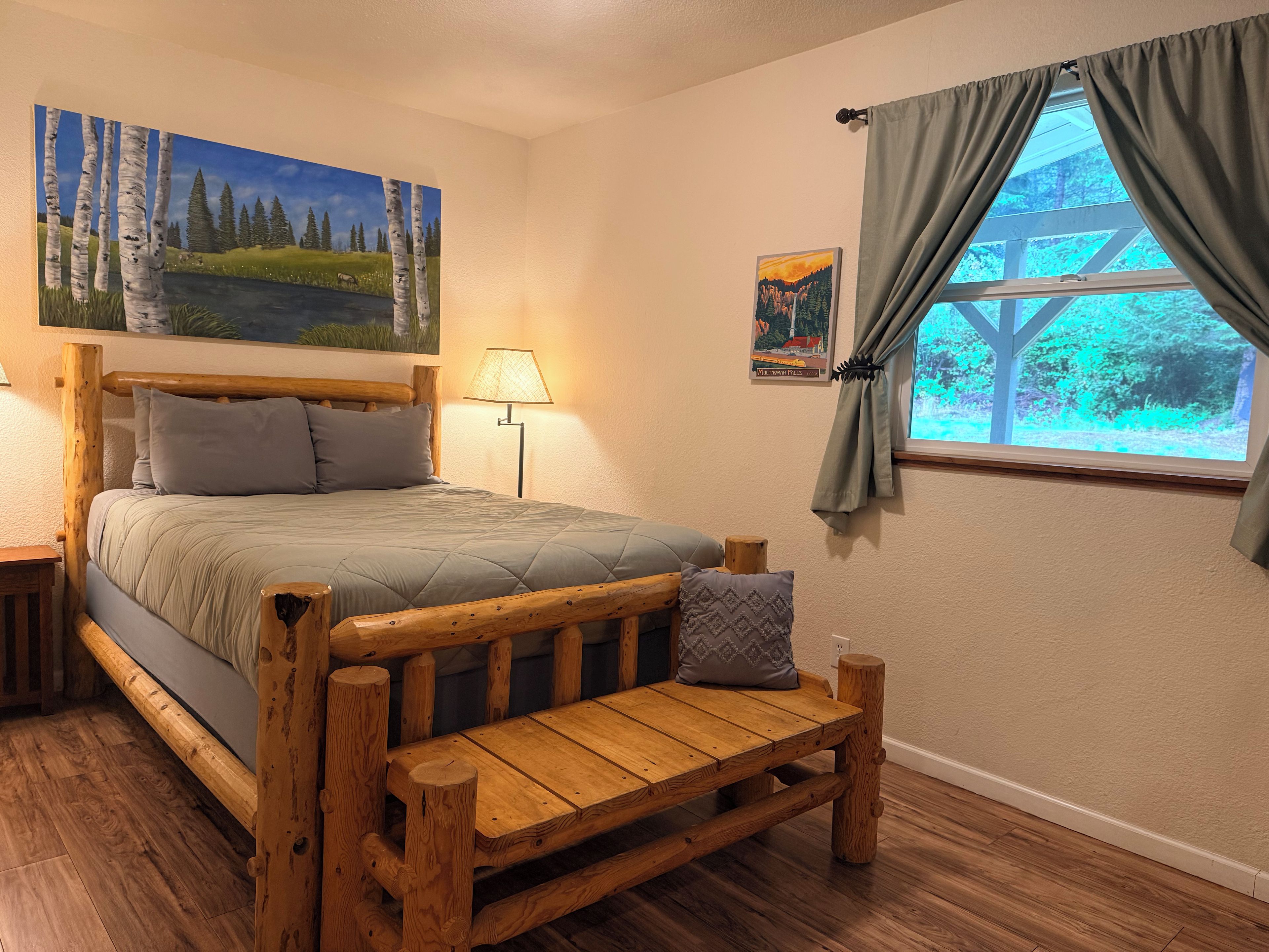 A bedroom with a wooden bed, a wooden bench, a painting of a landscape, a lamp, and a window with curtains.