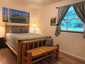A bedroom with a wooden bed, a wooden bench, a painting of a landscape, a lamp, and a window with curtains.