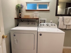 Un área de lavandería con dos lavadoras Whirlpool blancas, un estante de madera con un jarrón y una ventana arriba.