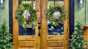 A wooden double door with glass panels, adorned with festive wreaths and surrounded by potted plants. - The Fir - 2 Bedroom Cabin (Broken Bow)