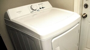 A front-loading white washer and dryer unit with a control panel, located in a laundry room with a wooden floor and a door in the background.