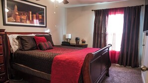 A bedroom with a large bed, a ceiling fan, a nightstand with a lamp, and a window with curtains.