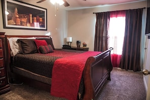 Quarto com uma cama grande, um ventilador de teto, mesa de cabeceira com abajur e janela com cortinas.