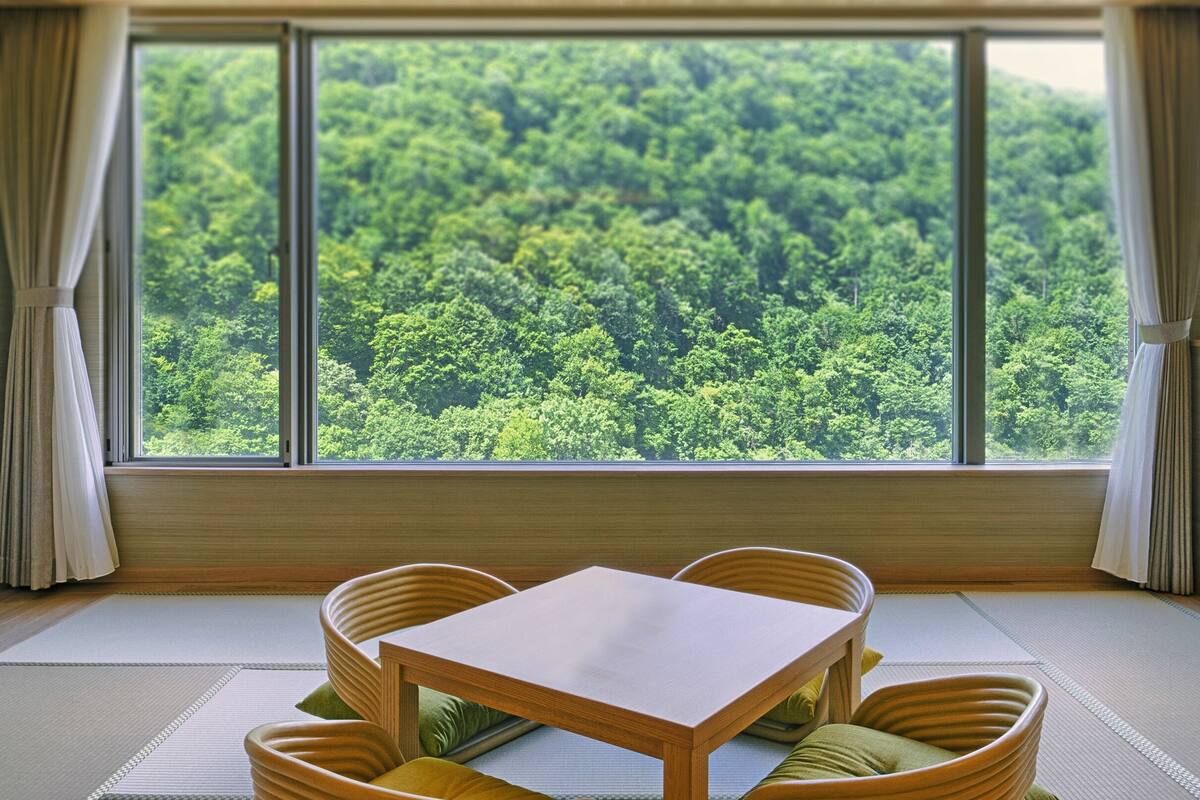 A room with tatami flooring, a low wooden table, and four tatami-style chairs. A large window offers a view of a dense forest.