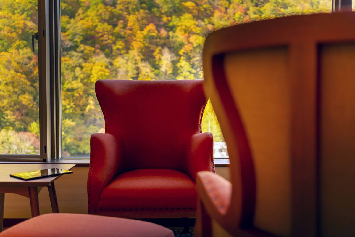 A red armchair by a window with a view of autumn foliage.