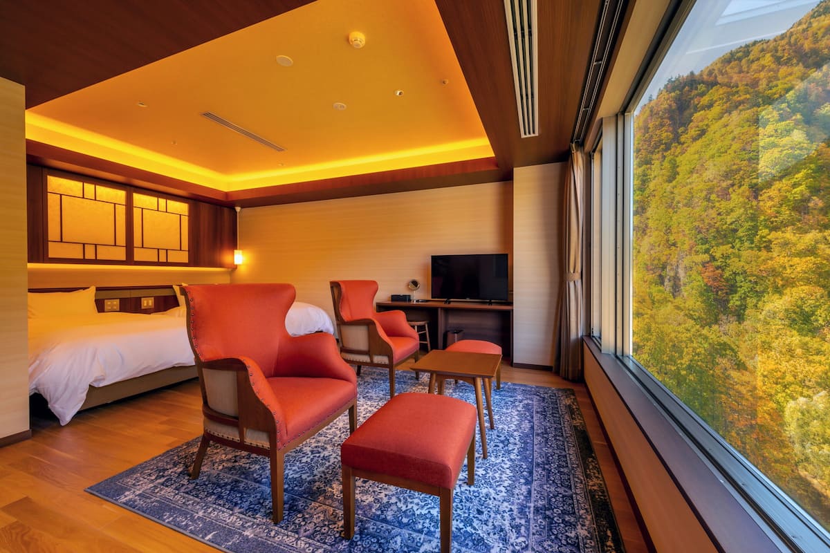 A modern hotel room with a bed, a seating area with a red chair and ottoman, a desk, and a television. The room has a large window offering a view of a forested hillside.