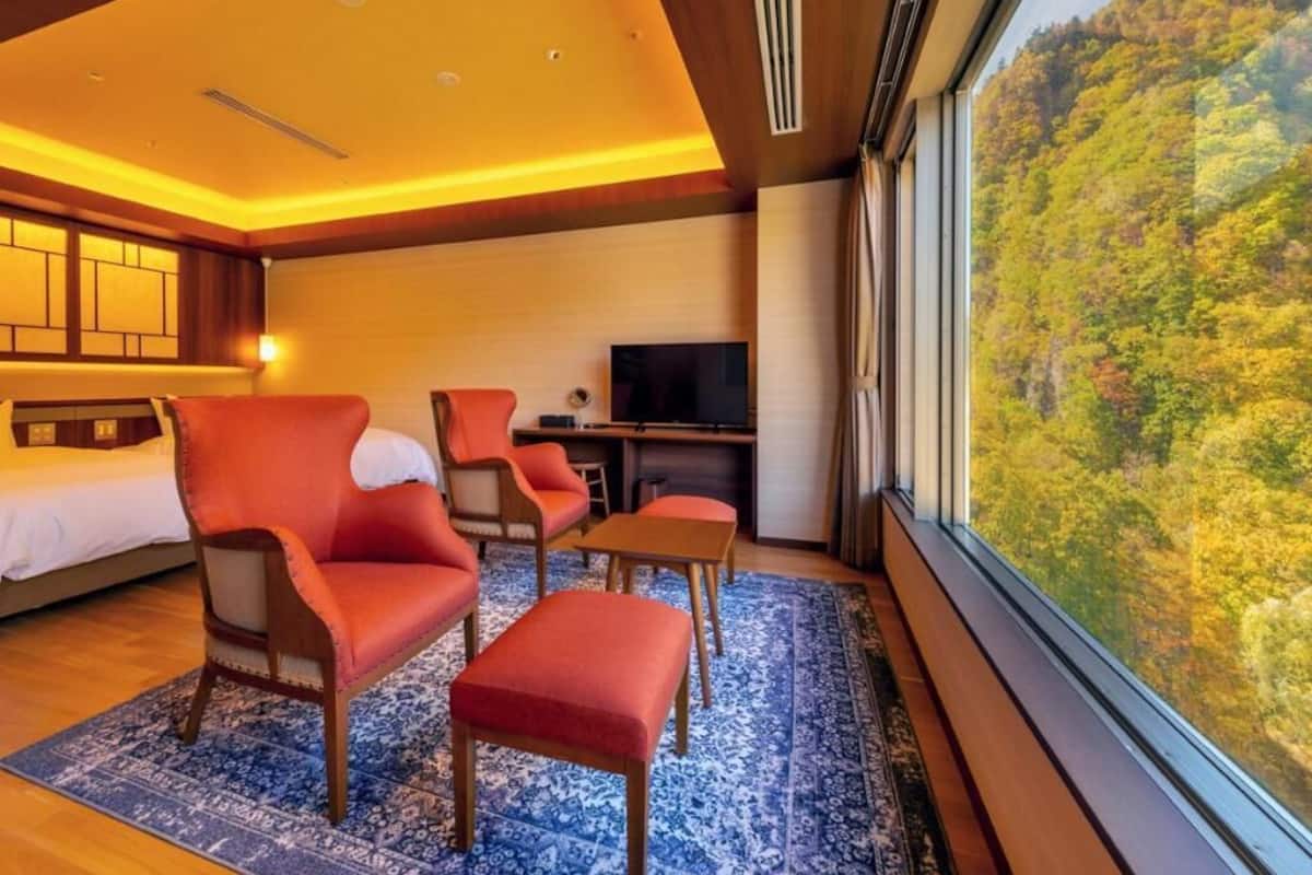 A hotel room with a bed, a TV, a seating area with chairs and a table, and a large window with a forest view.