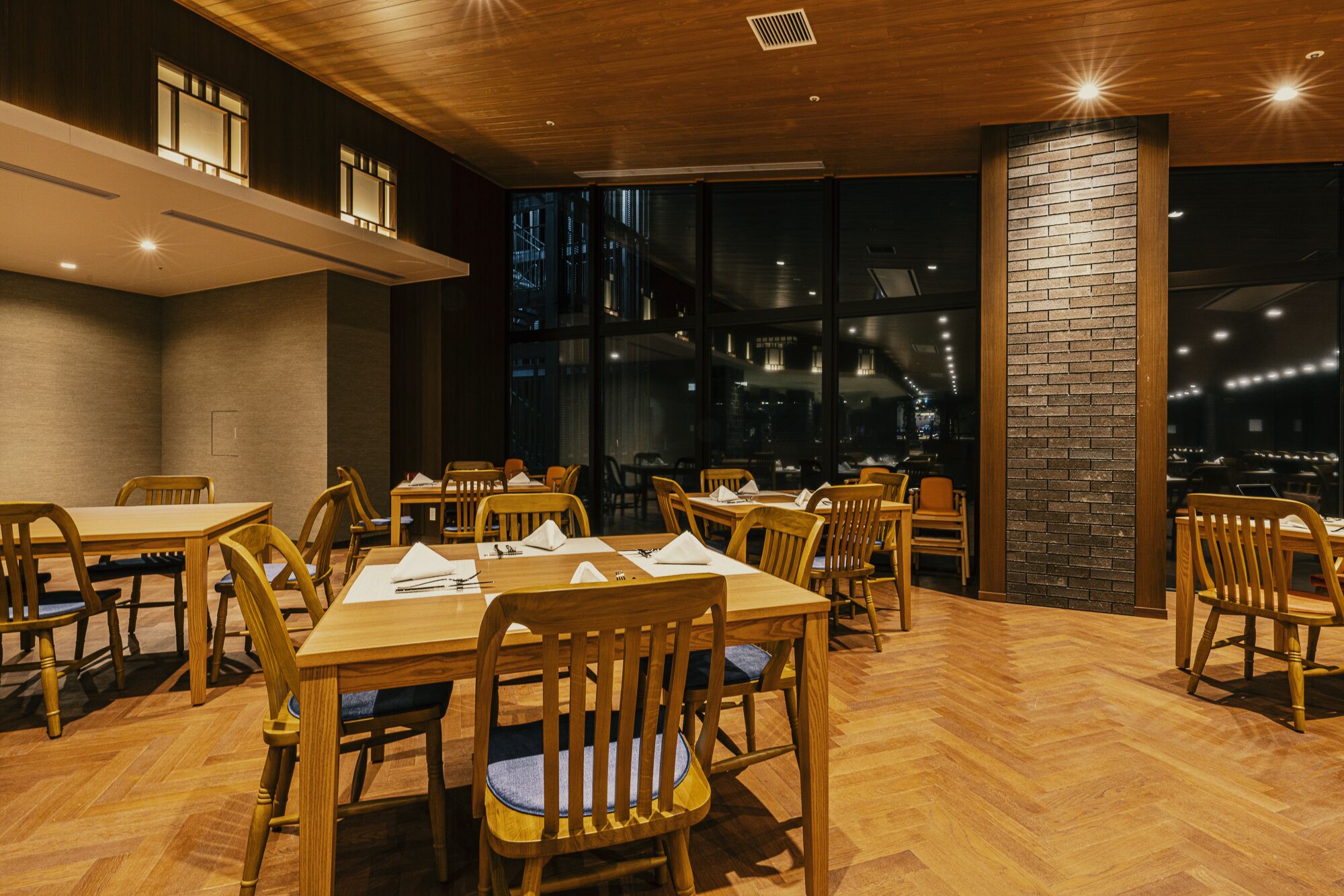 A restaurant with wooden tables and chairs, large windows, and a brick accent wall.
