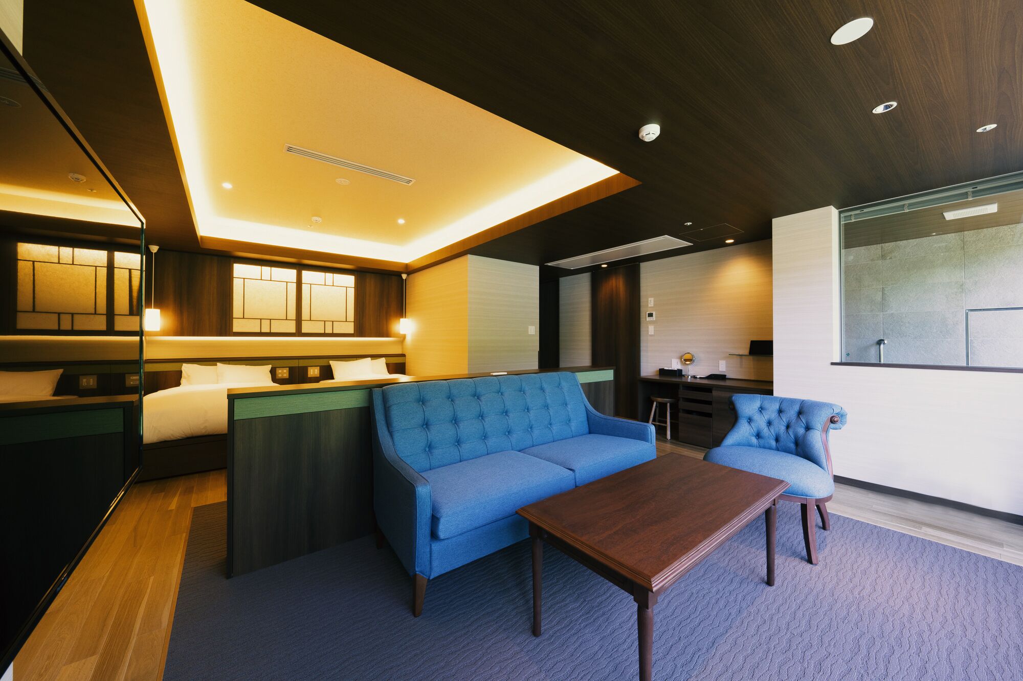 A modern hotel room with a blue sofa, a wooden coffee table, and a large window offering a view of the city.