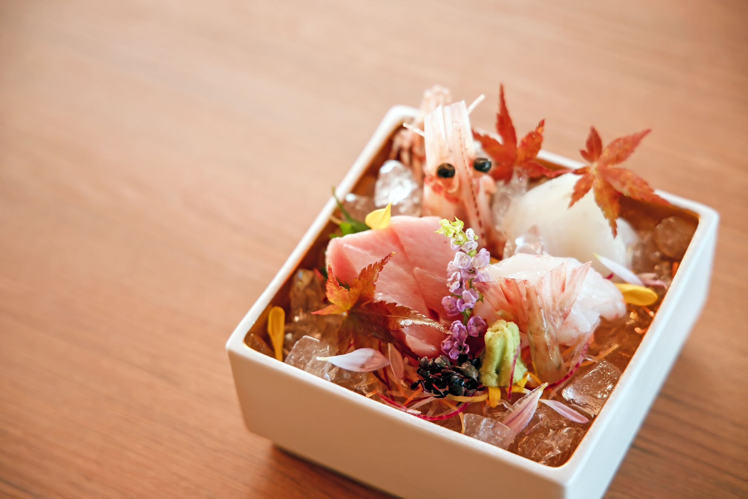 A square white bowl containing a seafood dish with ice, garnished with leaves and flowers.