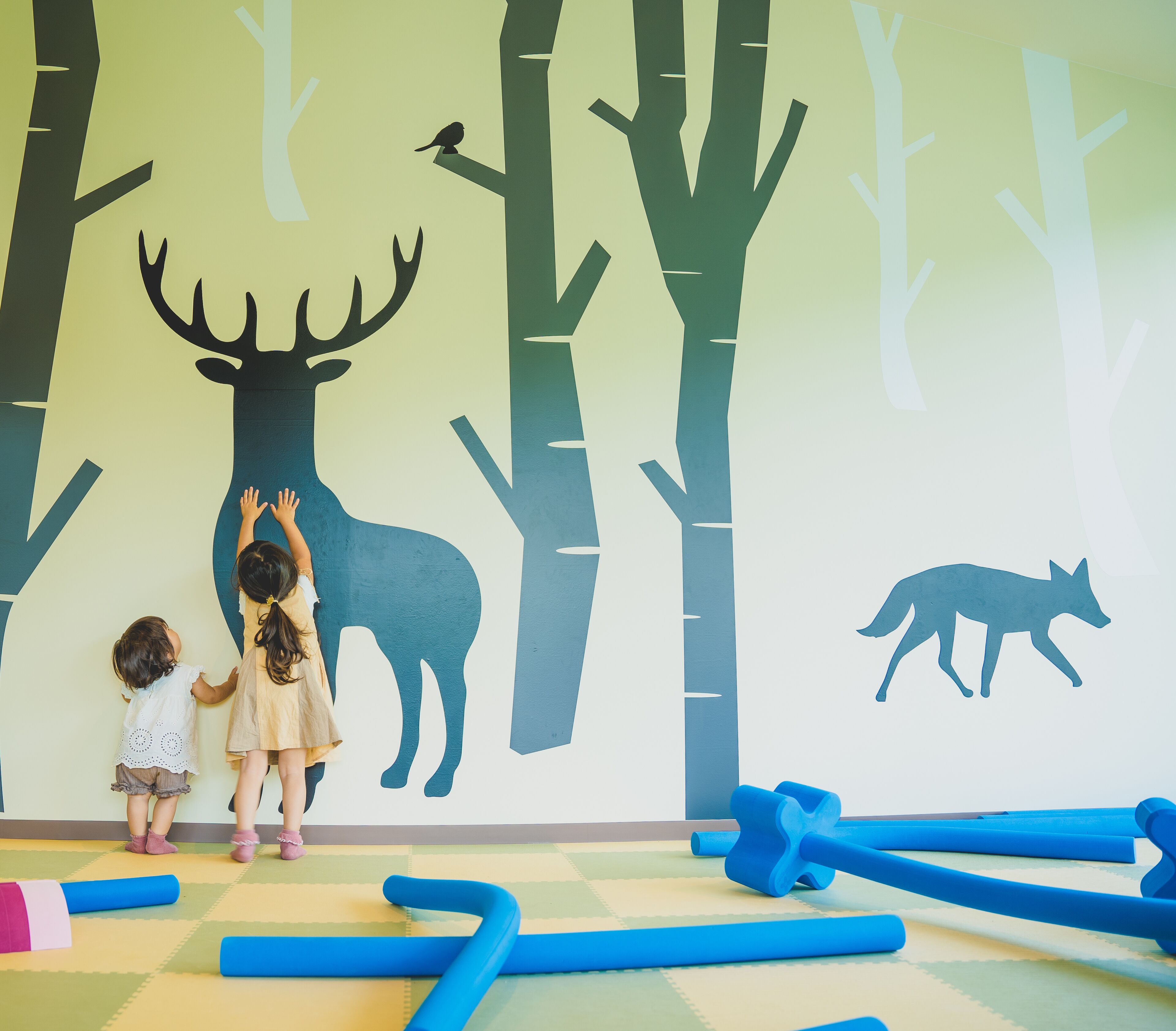 A room with a large wall mural of a forest scene, including a deer and a fox. There are blue foam mats on the floor.