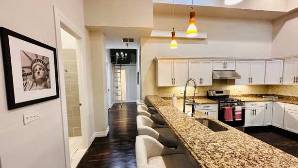 A modern kitchen with a long countertop, white cabinets, and a stainless steel stove. There is a hallway with a dark floor and a framed picture on the wall.