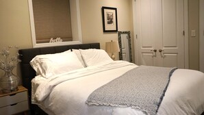 A neatly made bed with white linens and a gray blanket, a bedside table with a lamp, a framed picture on the wall, and a closed door.