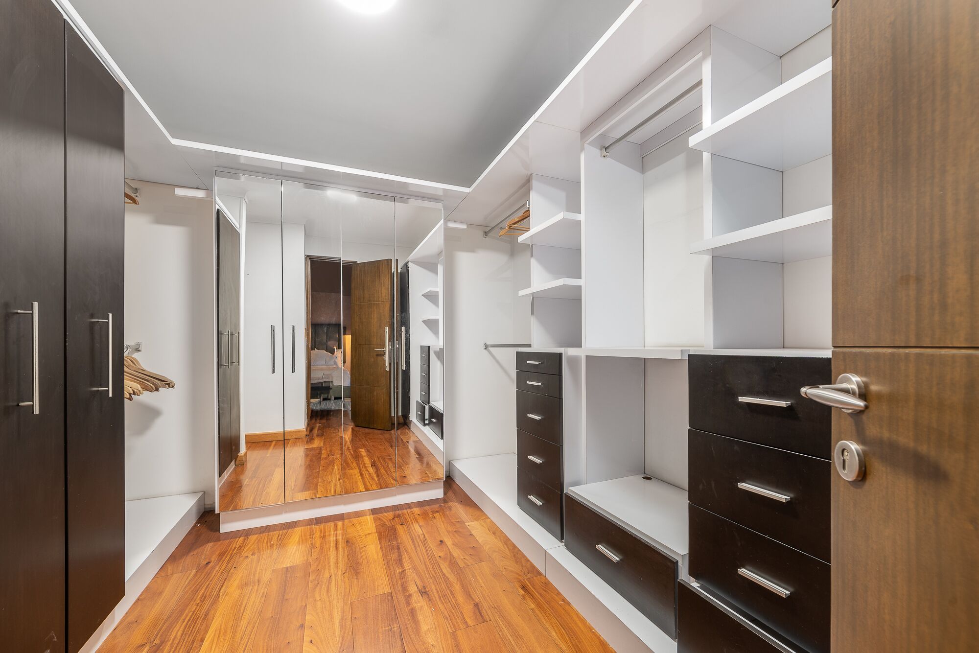 Un vestidor moderno con puertas espejadas, suelo de madera y armarios empotrados.