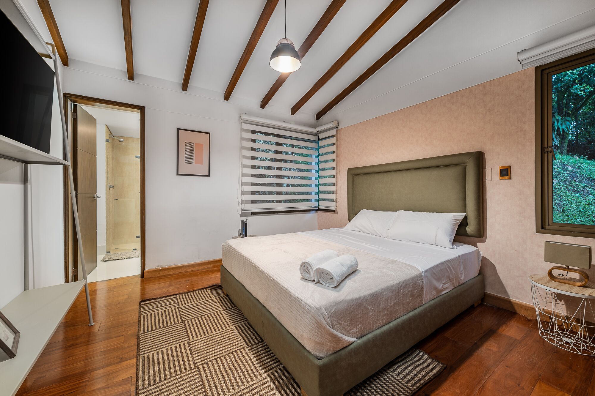 Une chambre moderne avec un grand lit, des poutres en bois au plafond, une fenêtre avec des stores et une vue sur la verdure.