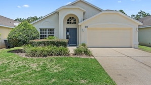 A single-story house with a garage, a front door, and a small front yard with bushes and a tree. - **Luxury 4 bedroom Villa w/ pool in golf community, mins away from DisneyWorld** (Davenport)