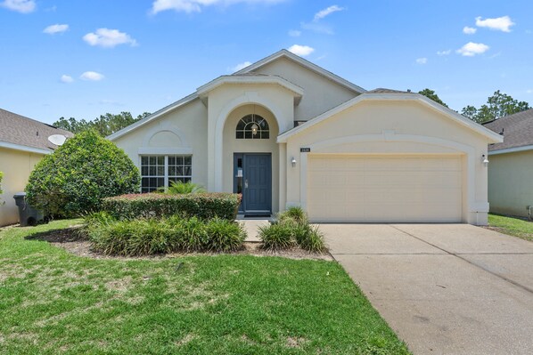 A single-story house with a garage, a front door, and a small front yard with bushes and a tree. - **Luxury 4 bedroom Villa w/ pool in golf community, mins away from DisneyWorld** (Davenport)