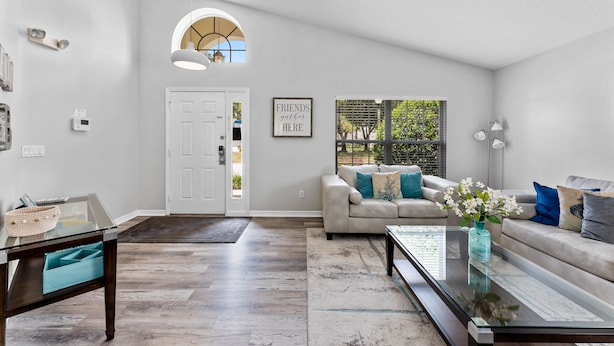 A spacious living room with a large window, a sofa, a coffee table, and a door leading to another room. - **Luxury 4 bedroom Villa w/ pool in golf community, mins away from DisneyWorld** (Davenport)