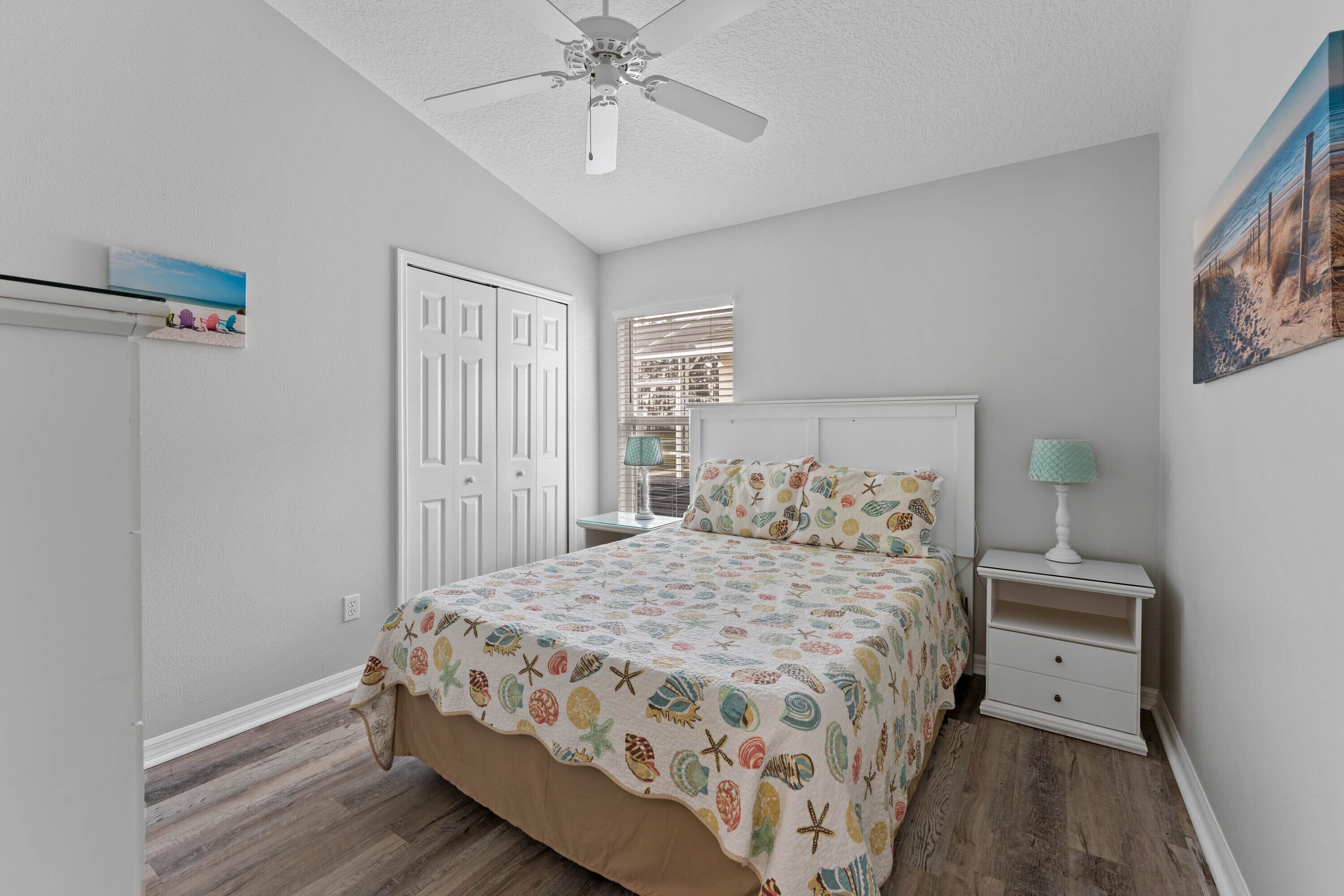Une chambre à coucher comprenant un lit, une table de chevet, un ventilateur de plafond, une fenêtre avec des stores et une porte.