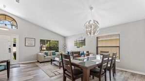 A spacious living room with a dining area, featuring a large glass-top table, dark wooden chairs, and a modern chandelier. - **Luxury 4 bedroom Villa w/ pool in golf community, mins away from DisneyWorld** (Davenport)